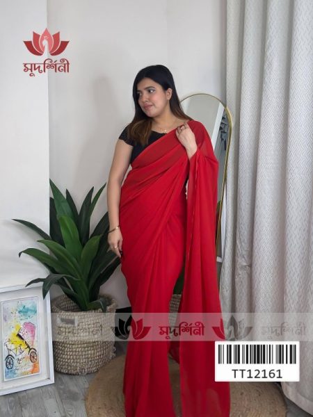 A woman wearing a plain red saree with a black blouse stands indoors near a mirror and houseplants. There is artwork on the floor and a white curtain in the background. Brand logos and a barcode are overlaid on the image.