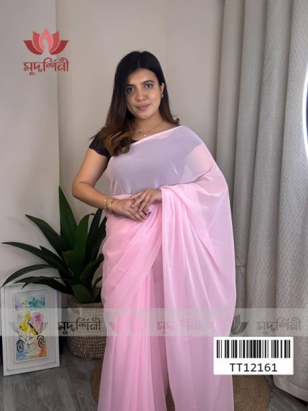 A woman in a light pink saree stands indoors near a potted plant and a light-colored curtain. She is smiling and wearing a black blouse. A framed artwork is on the floor beside her.