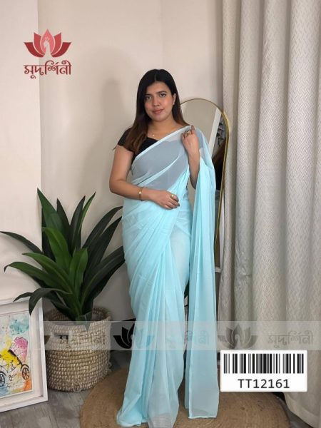 A woman wearing a light blue saree and black blouse stands indoors near a green plant and a floor mirror, with a beige curtain in the background and artwork on the floor.