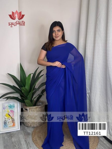 A woman in a royal blue saree with a black blouse stands indoors near a potted plant and a small table with books. The background includes a cream-colored curtain, wooden floor, and a “सुपर्णिका” logo on the wall.